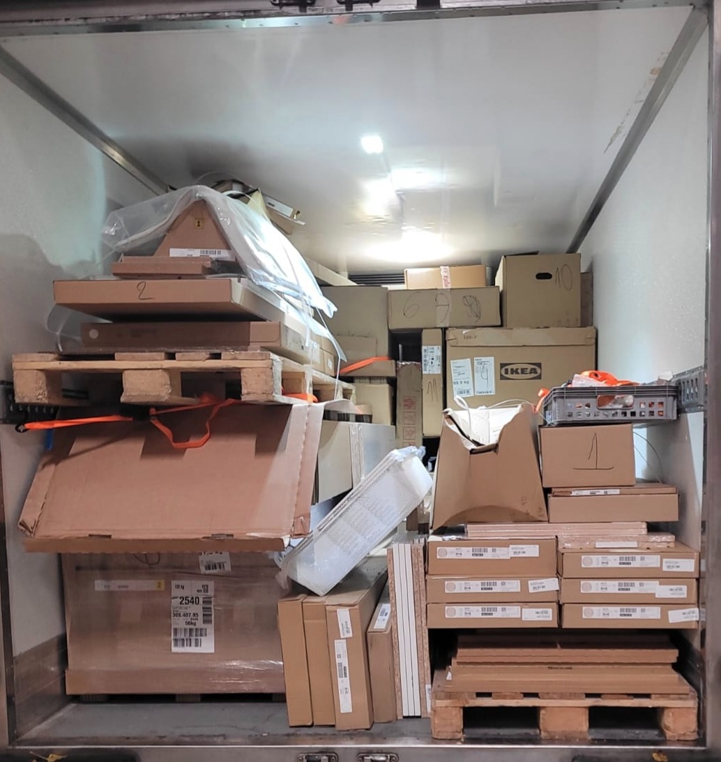 Wnętrze busa wypełnione kartonami, paletami i elementami mebli, gotowe do transportu. Widoczne oznaczenia IKEA na niektórych paczkach.