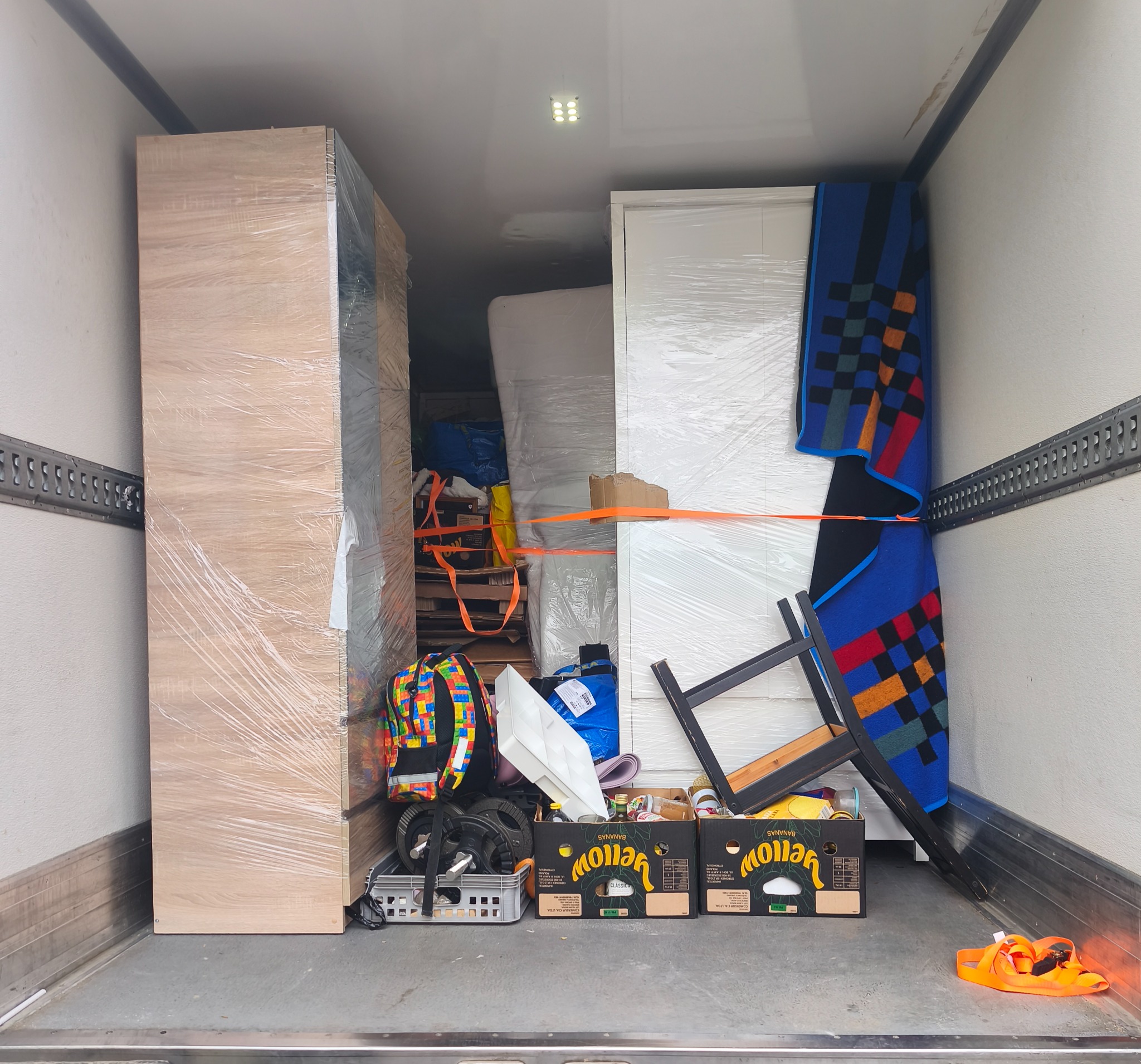 Wnętrze busa wypełnione meblami i kartonami zabezpieczonymi folią stretch i pasami transportowymi. Widoczne szafy, krzesło i kolorowy plecak.