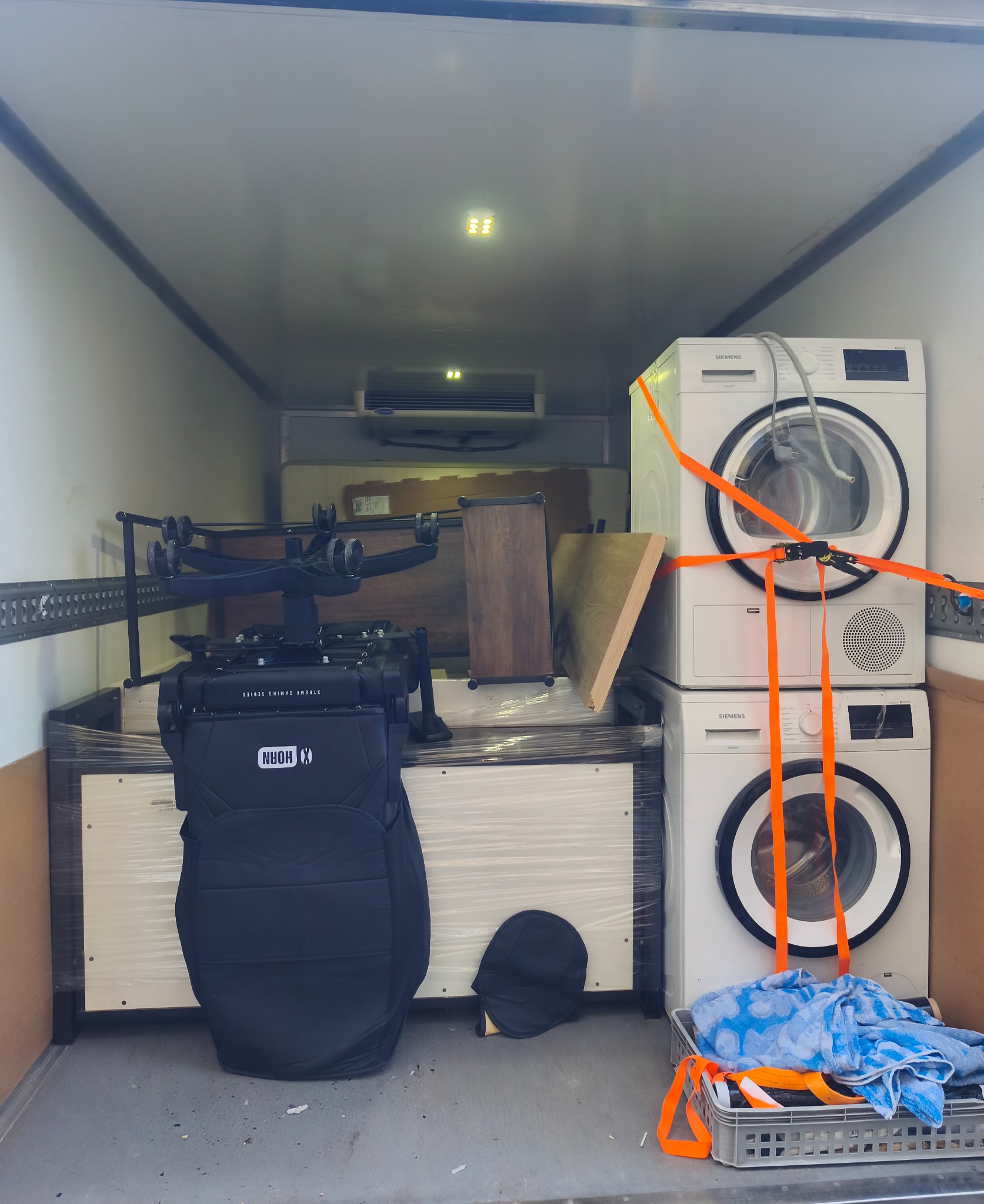 Wnętrze busa załadowane meblami (krzesło gamingowe, szafka) i sprzętem AGD (pralki, suszarka) zabezpieczonymi pomarańczowymi pasami transportowymi. Widoczne oświetlenie LED.