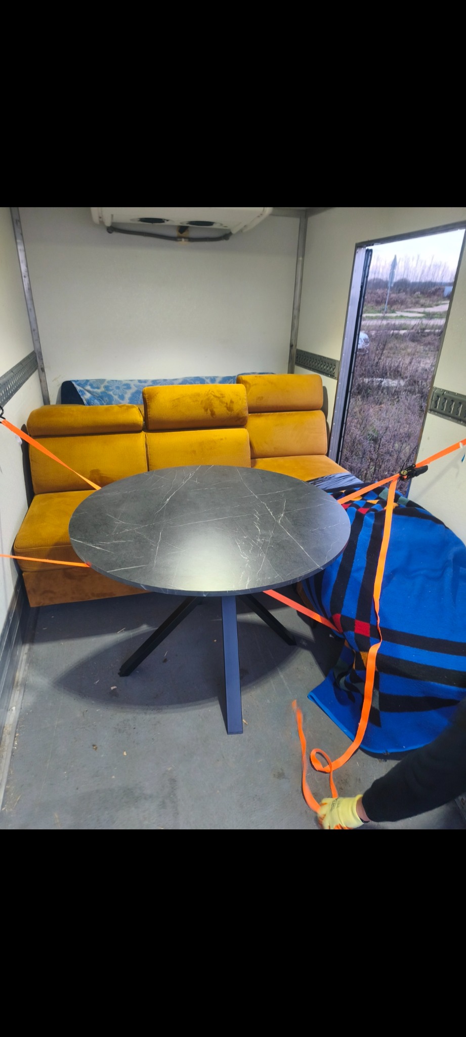 Wnętrze busa z zabezpieczonymi meblami: okrągły stół z blatem imitującym marmur, żółta sofa i niebieski koc, wszystko spięte pomarańczowymi pasami transportowymi.