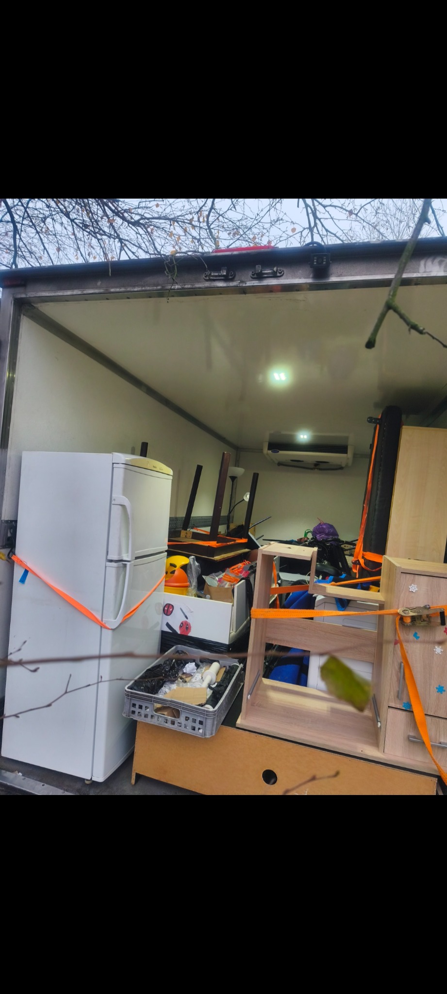 Wnętrze busa załadowane meblami i sprzętami AGD, zabezpieczonymi pomarańczowymi pasami transportowymi. Widoczne gałęzie drzew na zewnątrz pojazdu.