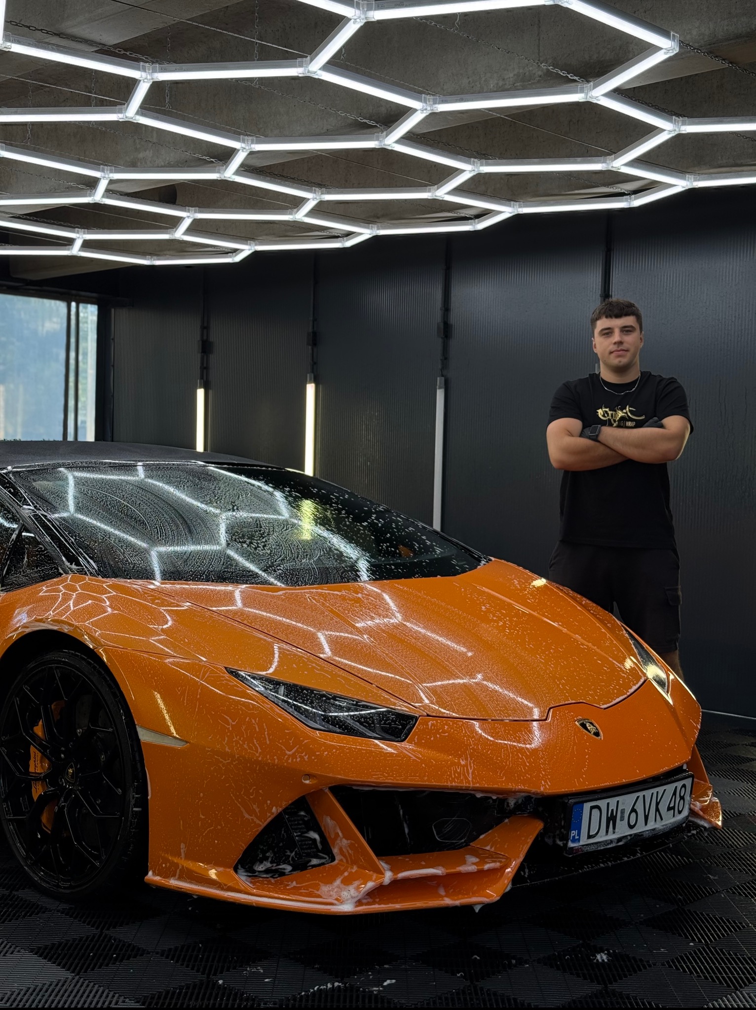 Pomarańczowe Lamborghini Huracan, pokryte pianą, myte w profesjonalnym studiu detailingu. Mężczyzna w czarnej koszulce stoi obok z założonymi rękami.