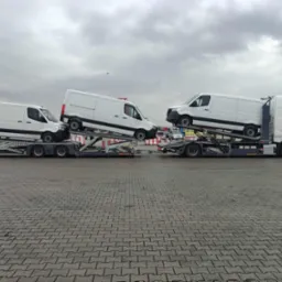 Dwie lawety z białymi busami na parkingu z kostki brukowej. Szara, pochmurna pogoda. Widoczne detale lawet i zabezpieczenia transportowe.