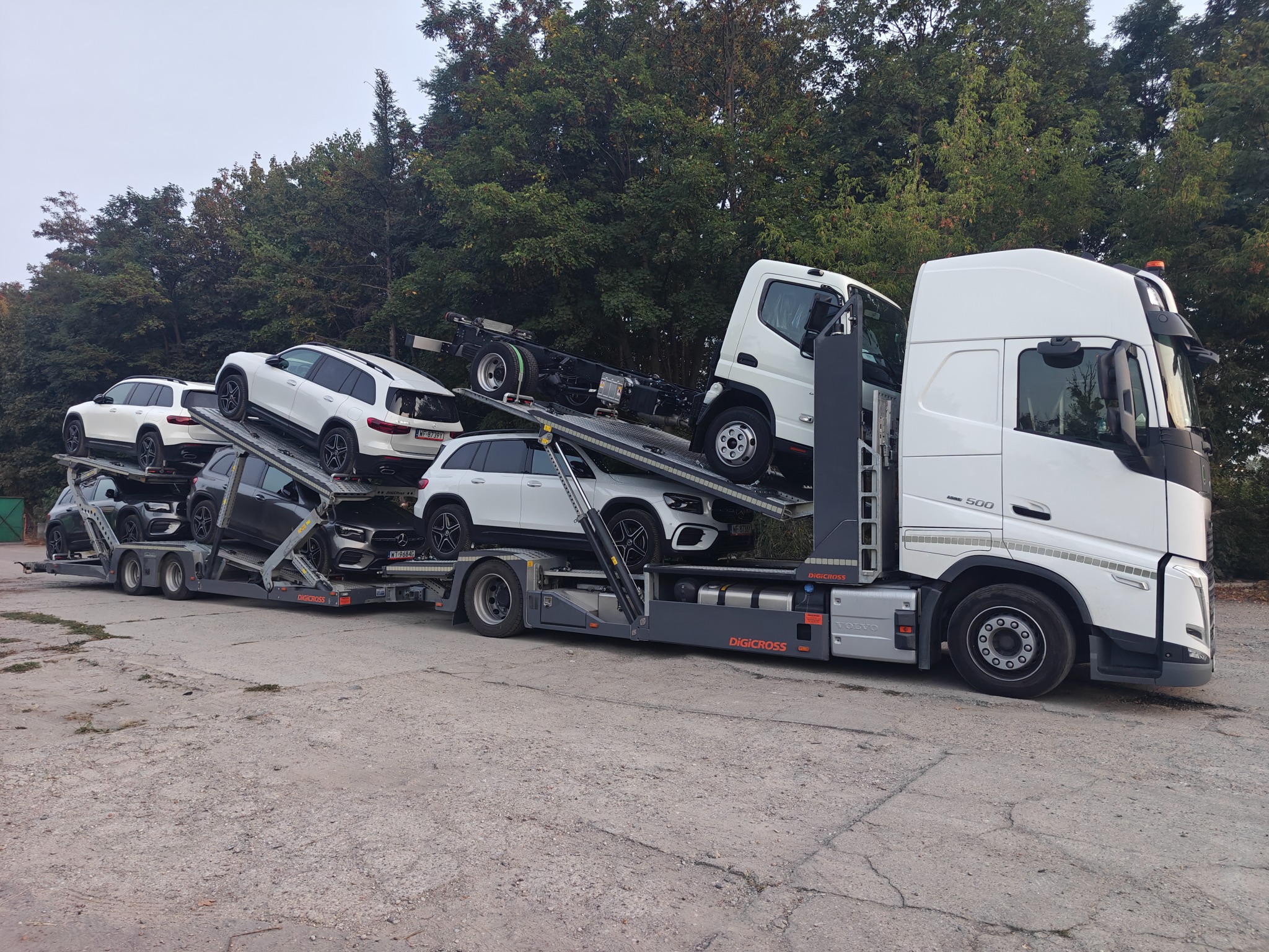 Laweta z załadowanymi samochodami osobowymi różnych marek i kolorów na tle zieleni, widok z boku. Ciągnik siodłowy Volvo z zabudową do przewozu aut.