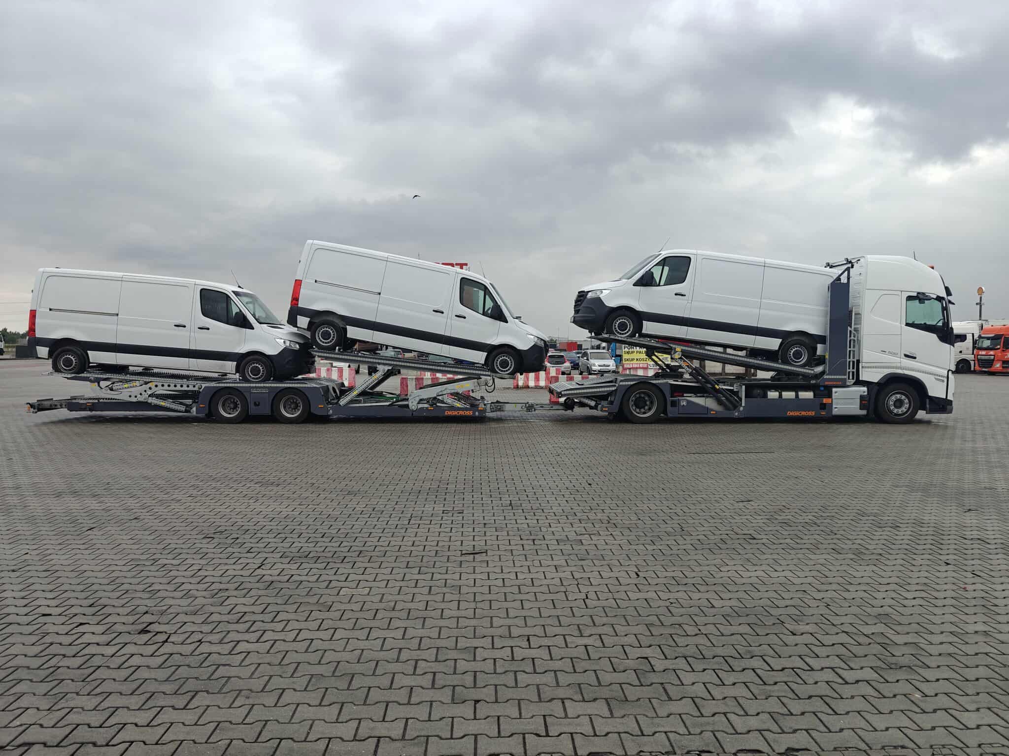 Dwie lawety z białymi busami na parkingu z kostki brukowej. Szara, pochmurna pogoda. Widoczne detale lawet i zabezpieczenia transportowe.