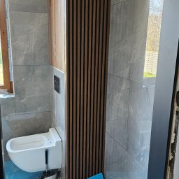 Marcin Stępień - Wykończona łazienka z szarymi płytkami imitującymi marmur, drewnianymi lamelami dekoracyjnymi i podwieszaną toaletą. Podłoga zabezpieczona folią ochronną.