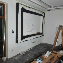 Marcin Stępień - Wnęka dekoracyjna z czarną ramą w trakcie montażu na białej ścianie. Widoczne przewody, drabina i narzędzia malarskie. Podłoga zabezpieczona folią.