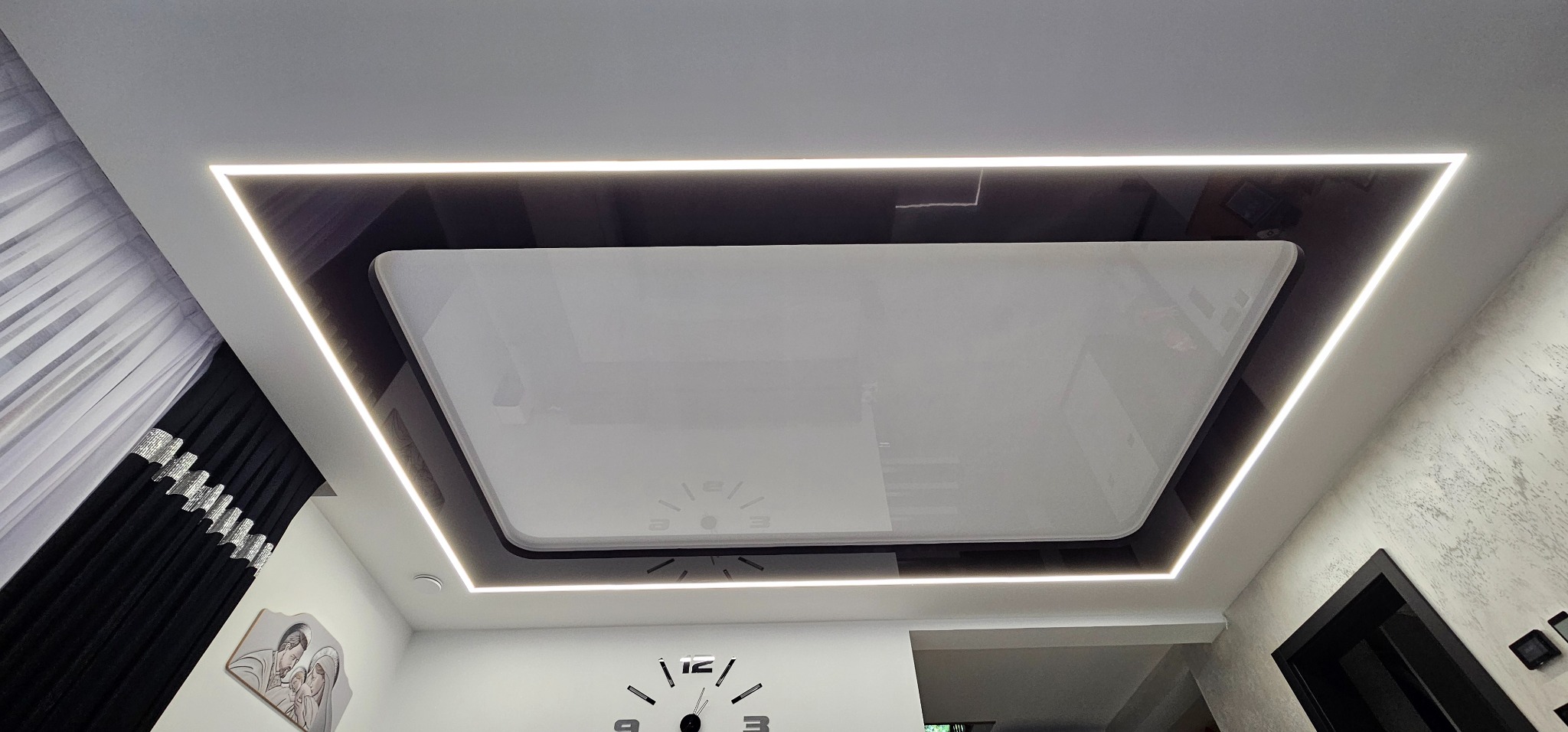 Nowoczesny sufit podwieszany z czarną wstawką i oświetleniem LED, widziany z dołu. Dekoracyjne zasłony i zegar na ścianie dopełniają wystrój wnętrza.