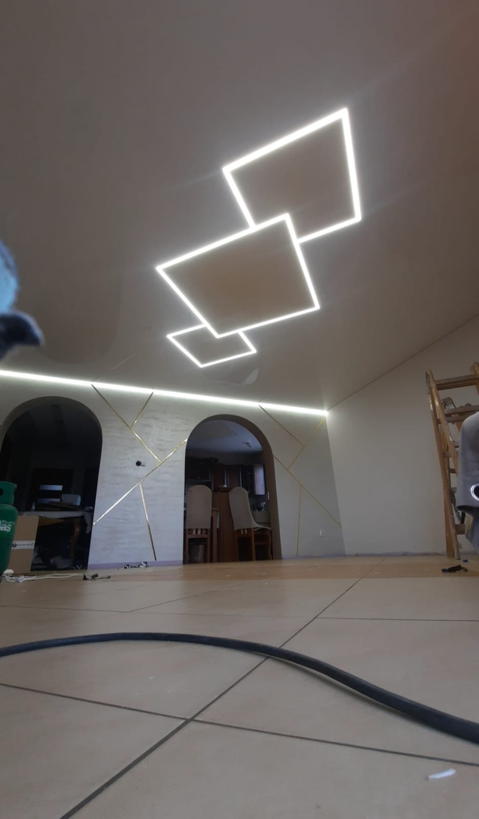 Nowoczesne oświetlenie sufitowe z kwadratowych paneli LED w jasnym wnętrzu z dekoracyjnymi architektonicznymi łukami i złotymi akcentami na ścianach.