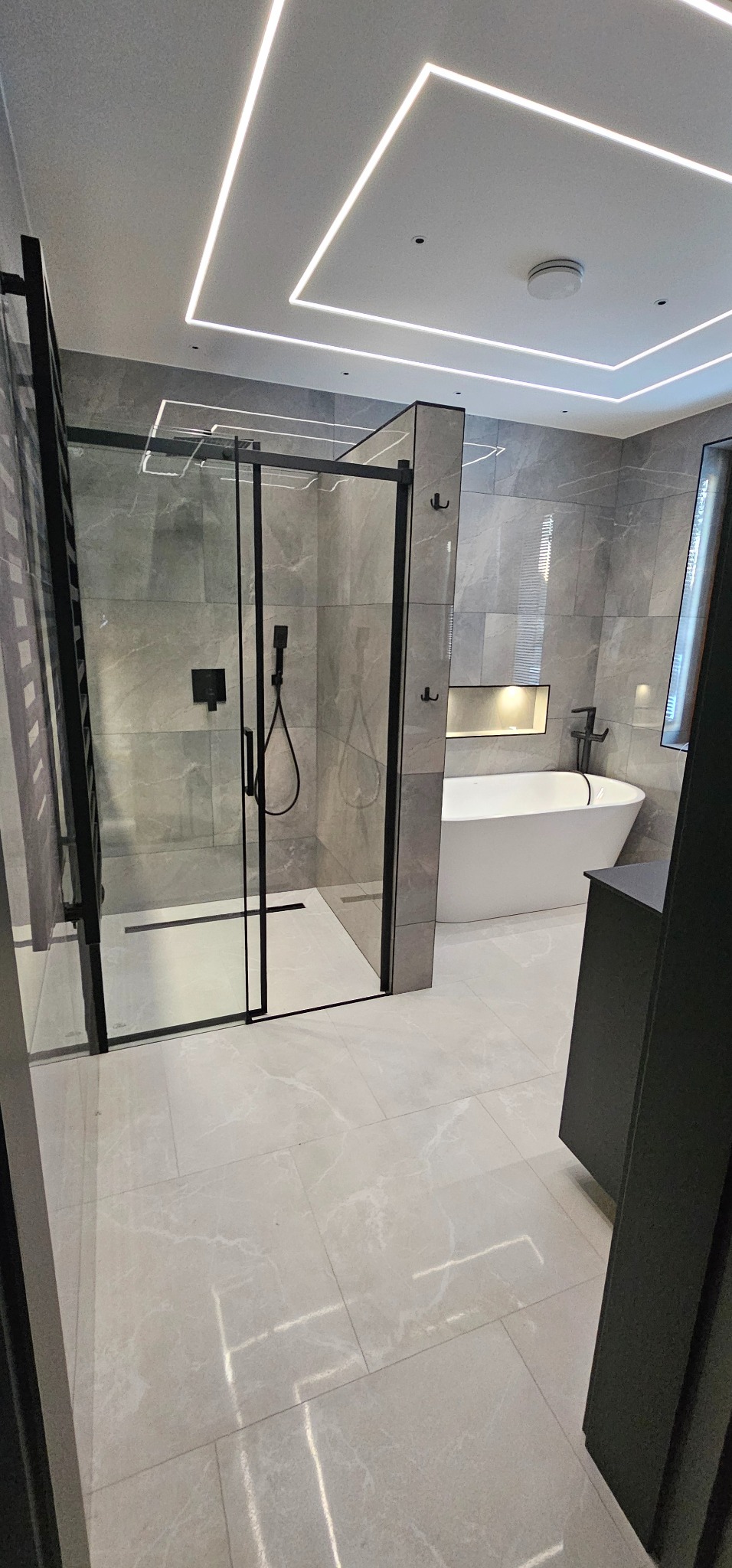 Nowoczesna łazienka z kabiną typu walk-in, szarymi płytkami i podświetlanym sufitem. Minimalistyczny design z czarnymi akcentami i wolnostojącą wanną.