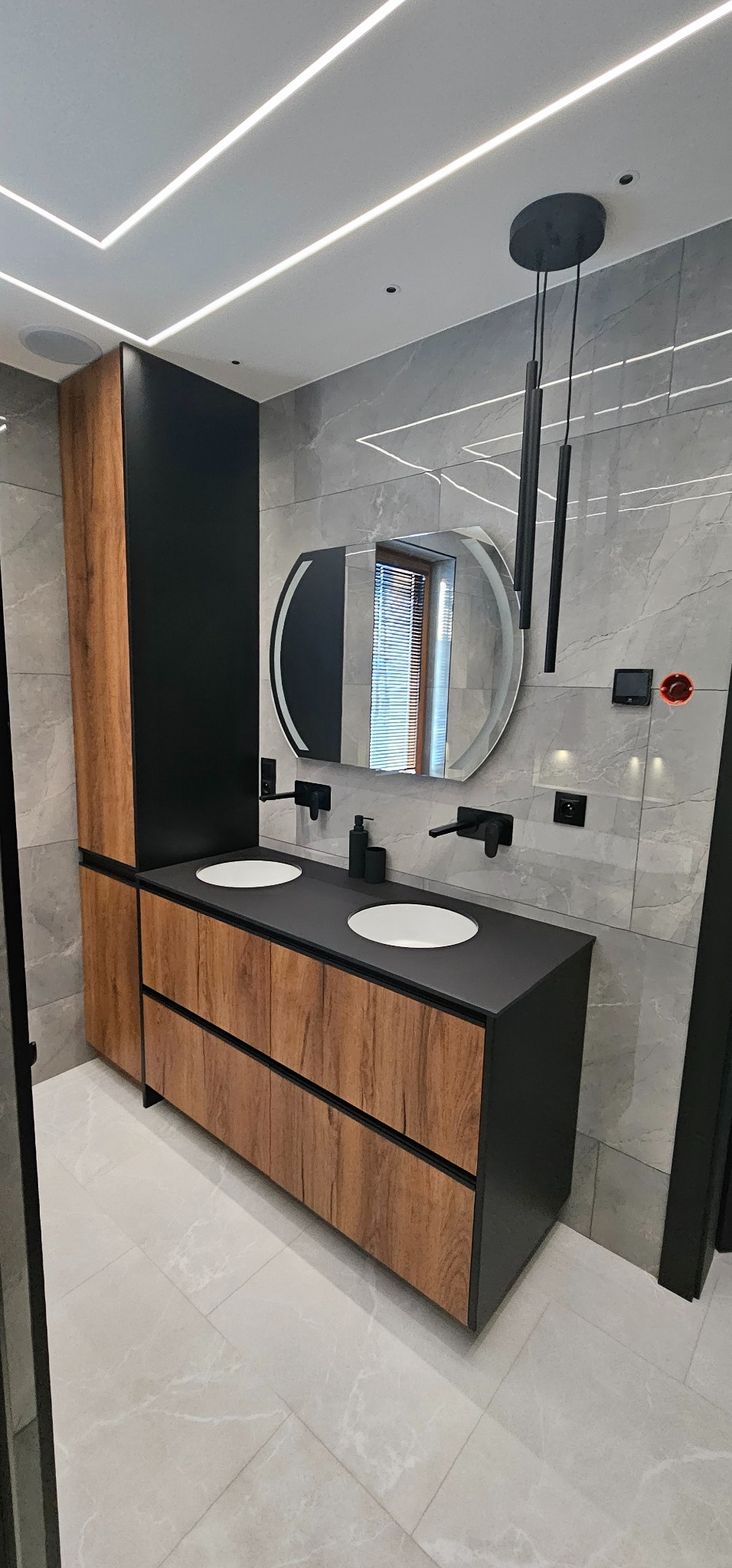 Nowoczesna łazienka z podwójną umywalką w szafce z drewnianym wykończeniem i czarnym blatem, oświetlona listwami LED w suficie i wiszącymi lampami.