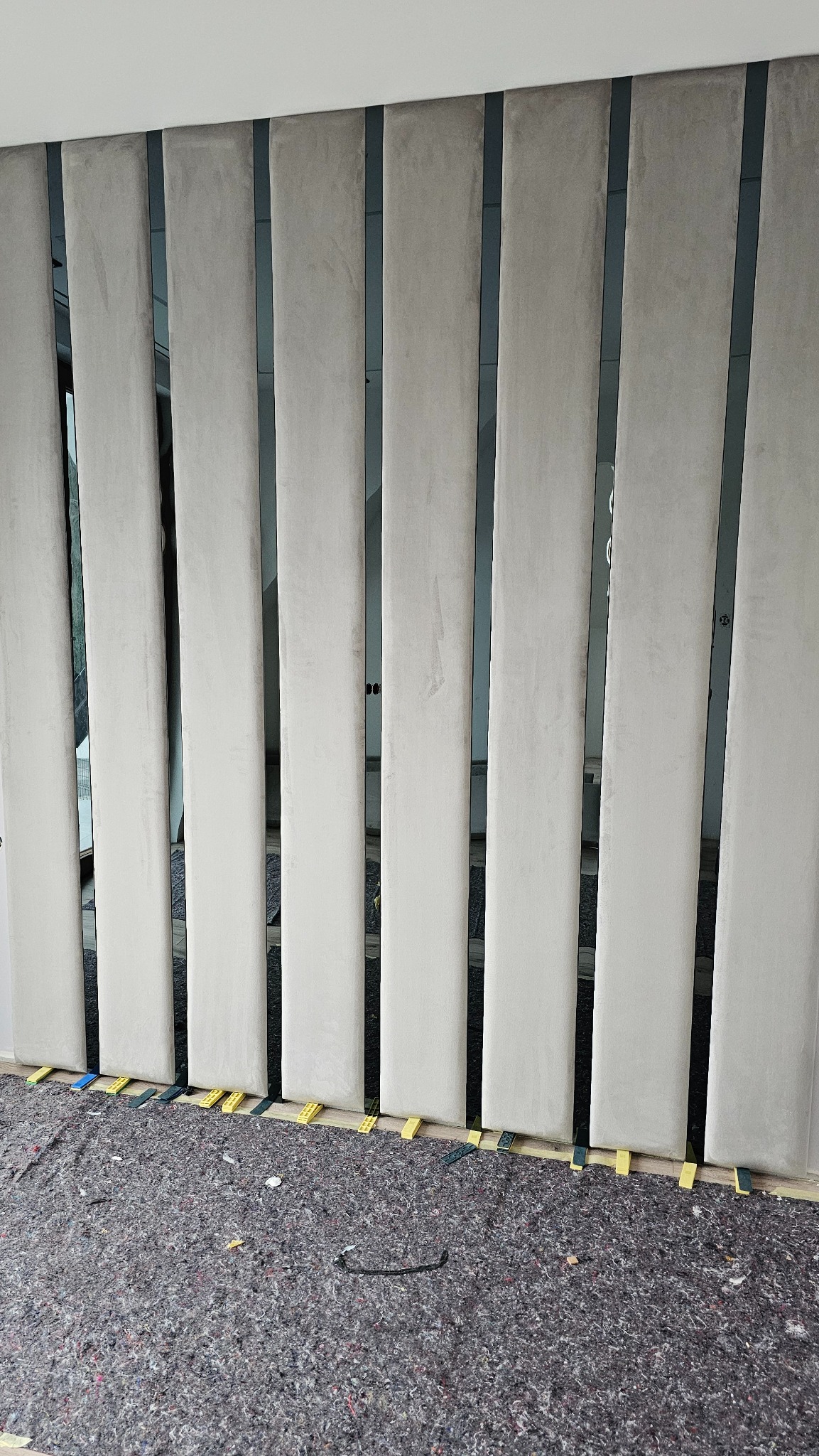 Montaż paneli ściennych z beżowej tkaniny, pionowe pasy oddzielone ciemnymi listwami. Na podłodze widoczne podkładki poziomujące i zabezpieczenie podłogi.