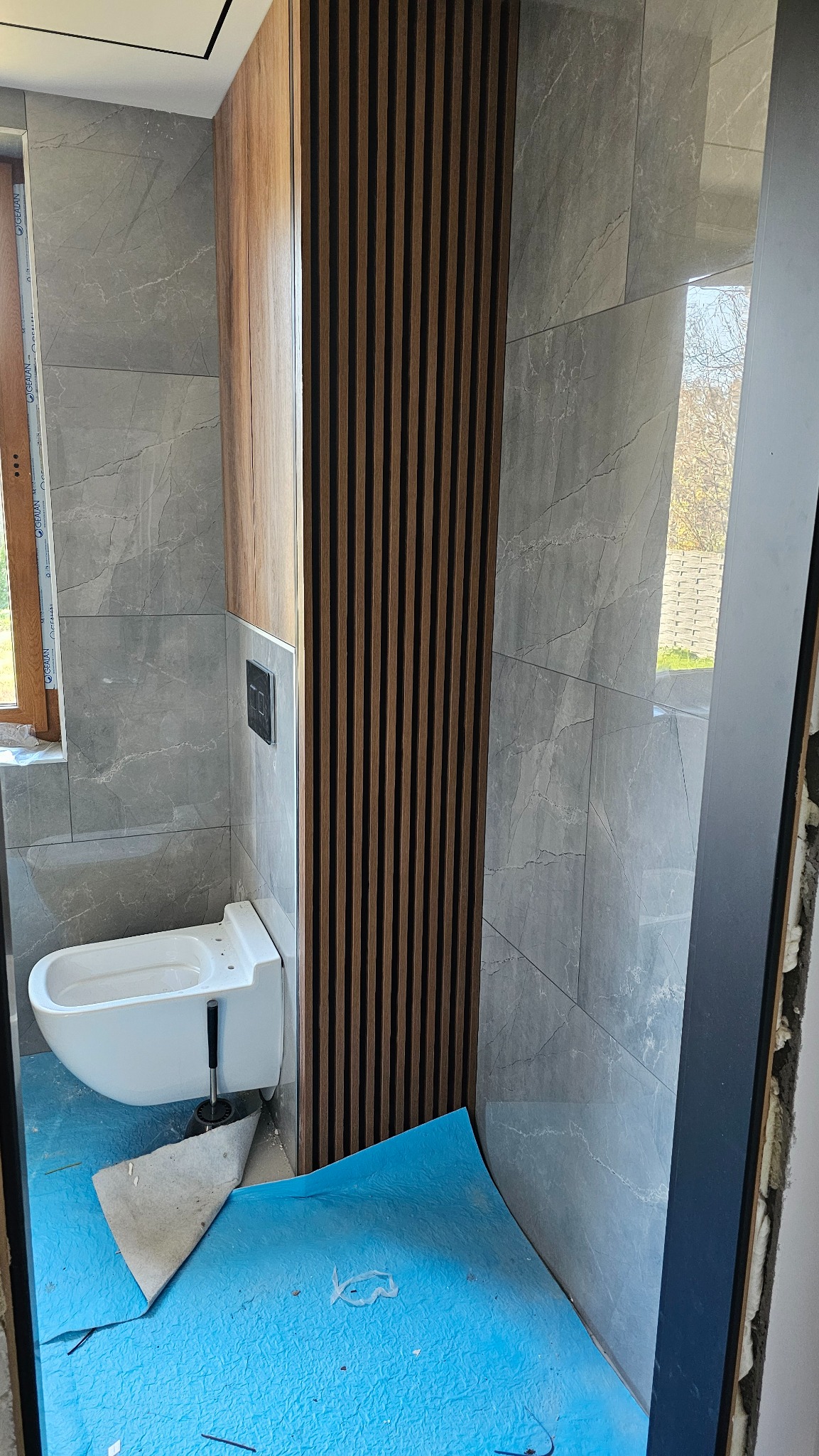 Wykończona łazienka z szarymi płytkami imitującymi marmur, drewnianymi lamelami dekoracyjnymi i podwieszaną toaletą. Podłoga zabezpieczona folią ochronną.