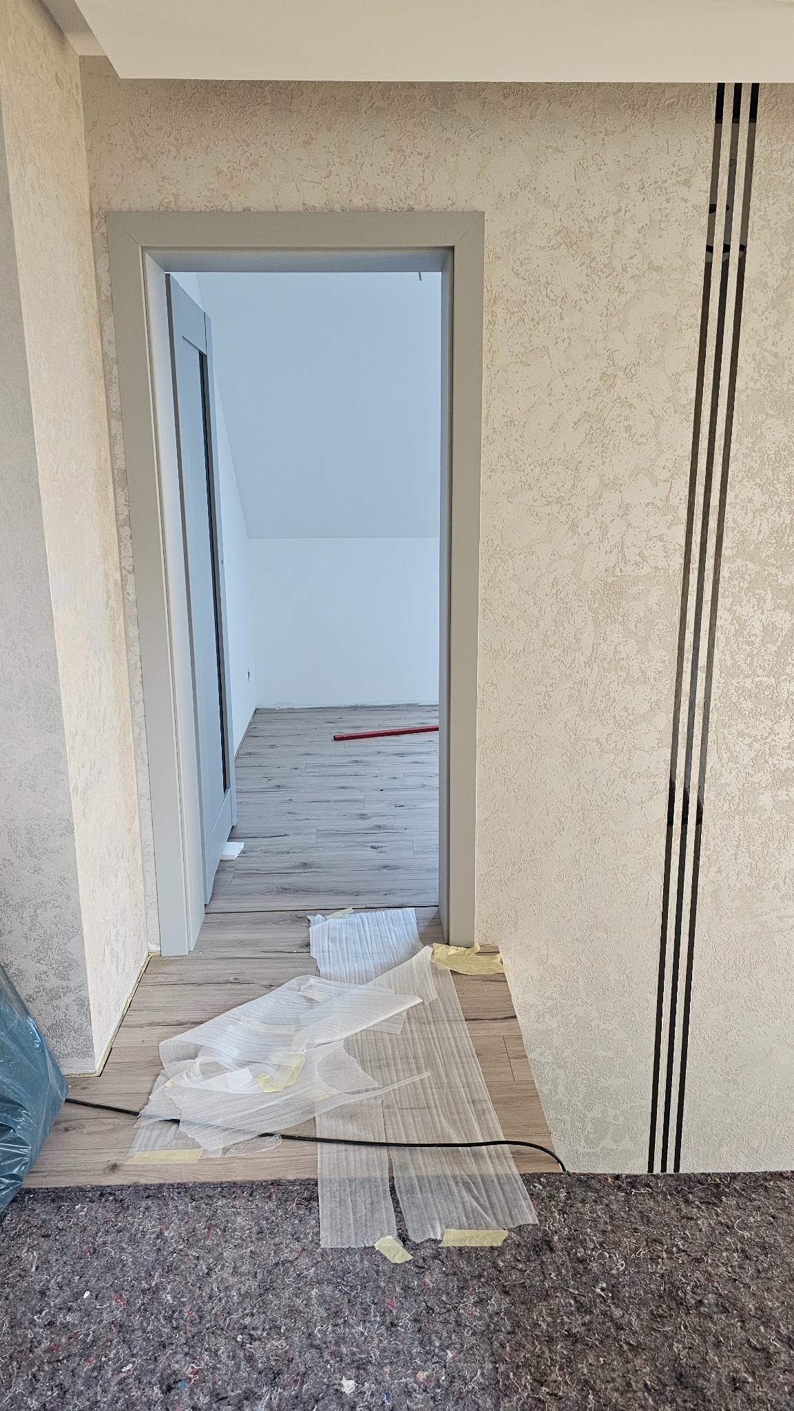 Remont pokoju: widok na świeżo położone panele podłogowe przez szare futryny drzwiowe. Na ścianie dekoracyjna tapeta z czarnymi paskami.