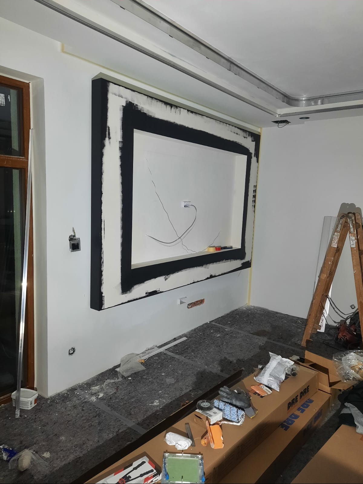 Wnęka dekoracyjna z czarną ramą w trakcie montażu na białej ścianie. Widoczne przewody, drabina i narzędzia malarskie. Podłoga zabezpieczona folią.