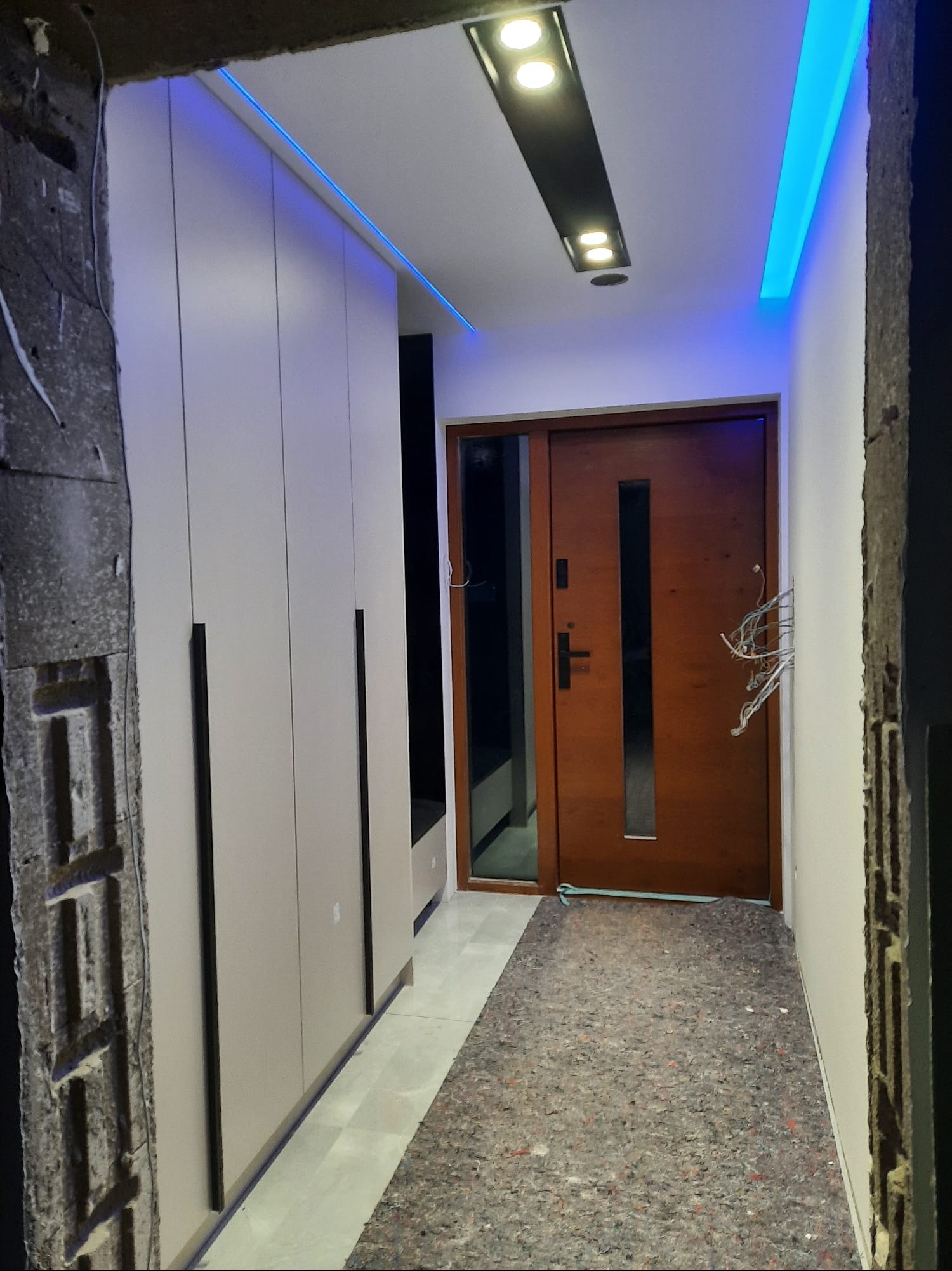 Nowoczesny przedpokój z szafą wnękową, oświetleniem LED i drewnianymi drzwiami wejściowymi. Fragment ściany w stanie surowym, widoczne przewody elektryczne.