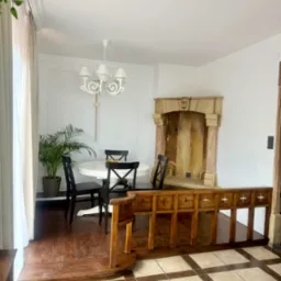 Jasne wnętrze z okrągłym stołem i czarnymi krzesłami, dekoracyjnym kominkiem i drewnianą balustradą. Widoczna roślina doniczkowa i biały żyrandol.