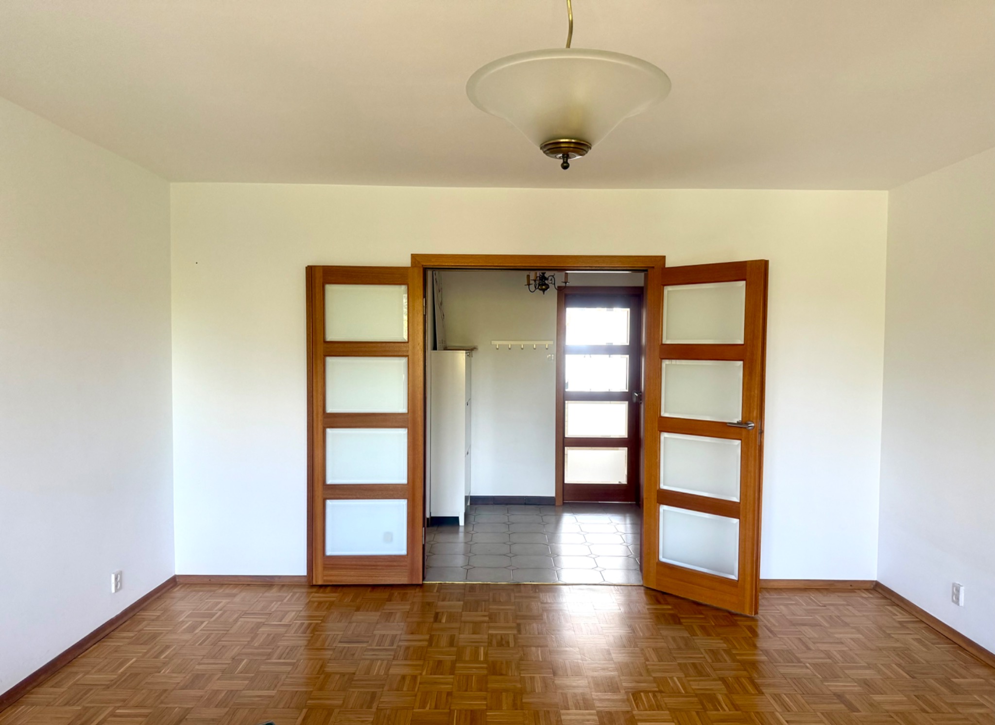 Jasne wnętrze mieszkania z parkietem i rozsuwanymi drzwiami z matowego szkła, widok na pomieszczenie gospodarcze. Lampa sufitowa w stylu retro.