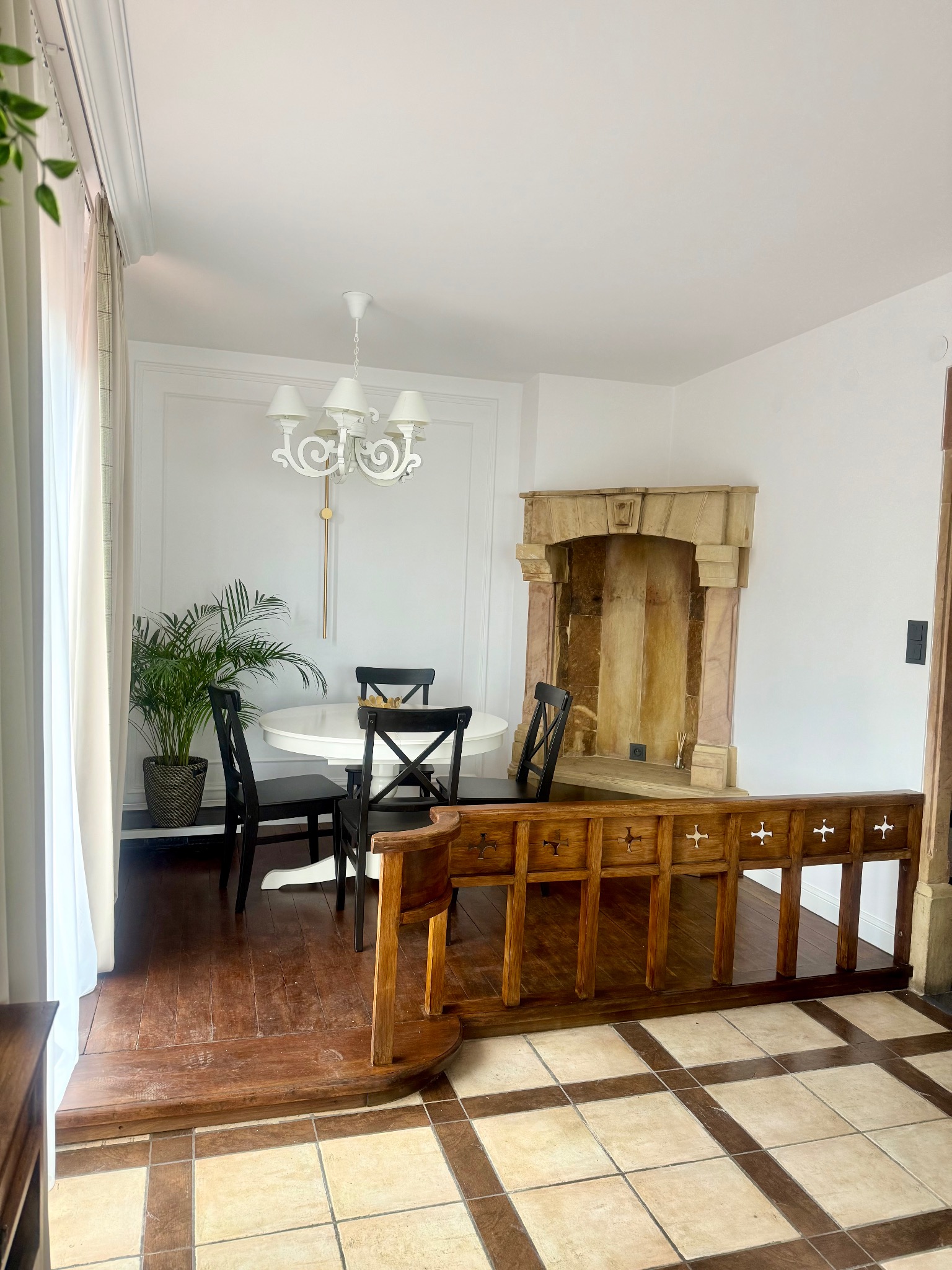 Jasne wnętrze z okrągłym stołem i czarnymi krzesłami, dekoracyjnym kominkiem i drewnianą balustradą. Widoczna roślina doniczkowa i biały żyrandol.