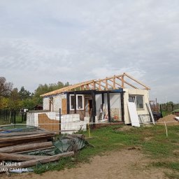 Emil Myjak - Budowa domu w trakcie: widoczna drewniana więźba dachowa na surowym budynku, teren budowy z materiałami i zielenią w tle.