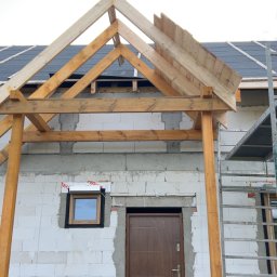 Emil Myjak - Budowa zadaszenia nad wejściem do domu z białego bloczka. Widoczna drewniana konstrukcja dachu i fragment pokrycia. Na ścianie okno i brązowe drzwi.