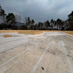 Emil Myjak - Montaż membrany dachowej Rothoblaas na drewnianej konstrukcji. Widok z góry na płaską powierzchnię z widocznymi drzewami i budynkiem w tle.