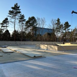 Emil Myjak - Wczesny etap budowy domu z drewnianych paneli na fundamencie, widoczny żuraw budowlany, w tle las i budynek biurowy. Słoneczny dzień.