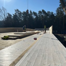 Emil Myjak - Montaż drewnianej konstrukcji dachu na budowie, widoczni pracownicy i dźwig. Elementy konstrukcyjne ułożone na przygotowanej powierzchni, las w tle, słoneczny dzień.