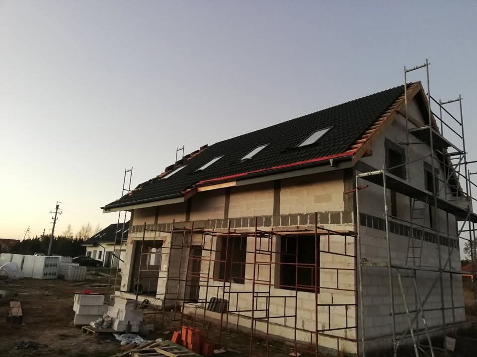 Dom w trakcie budowy z czarnym dachem z oknami połaciowymi i czerwonymi elementami wykończeniowymi, otoczony rusztowaniami. Widoczne materiały budowlane.