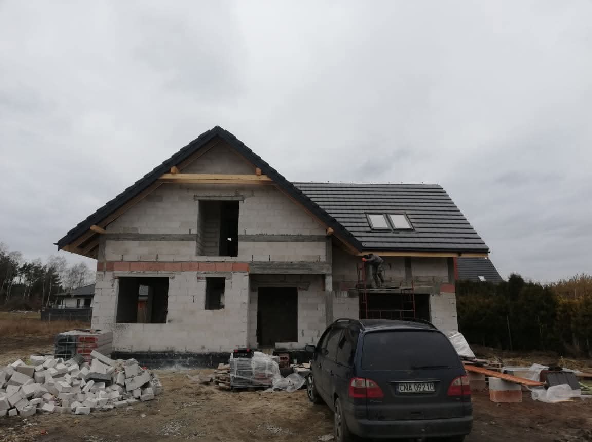 Budowa domu jednorodzinnego w trakcie krycia dachu dachówką. Widoczny pracownik na rusztowaniu, częściowo pokryty dach z oknami dachowymi i sterta dachówek.