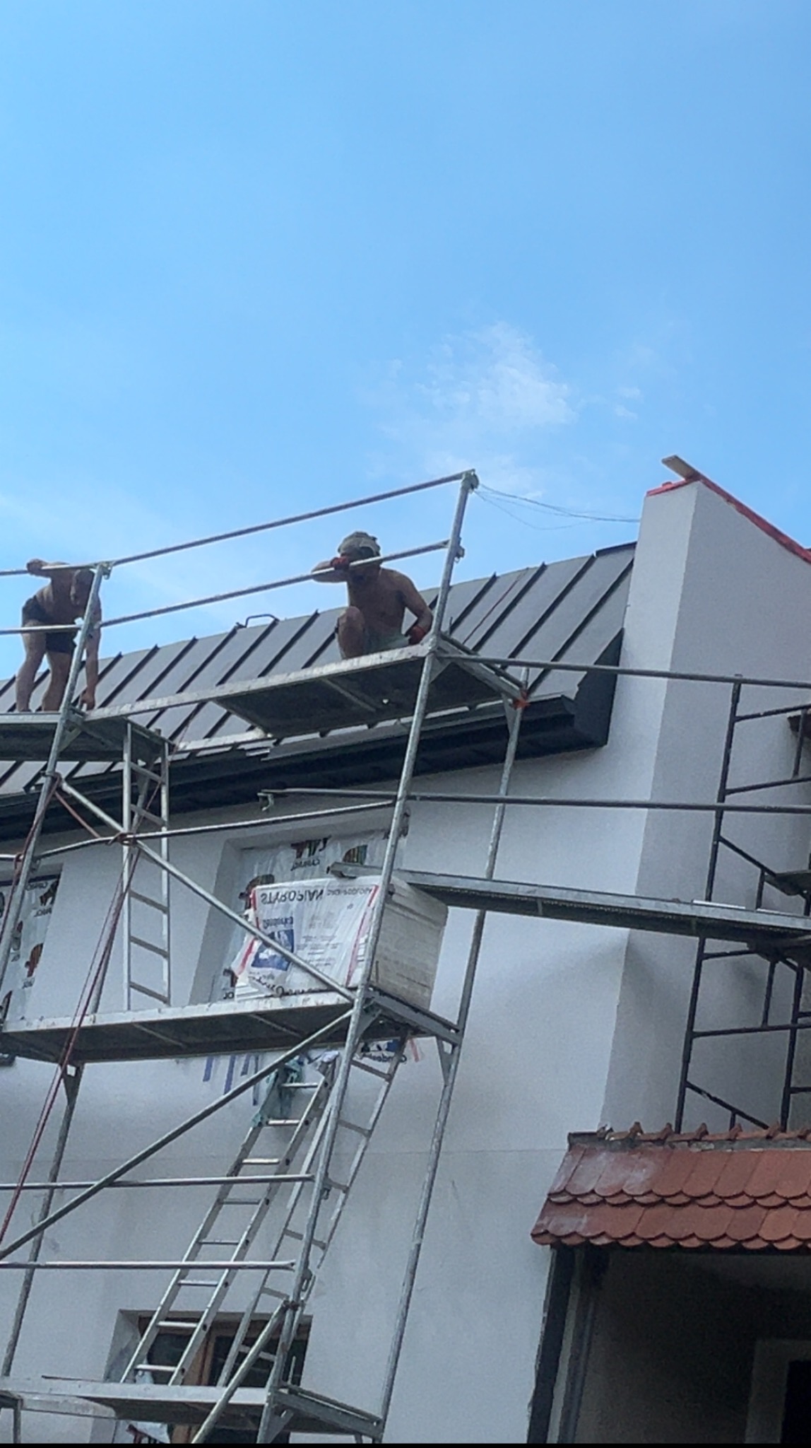 Praca na dachu na rąbek: dwaj dekarze na rusztowaniu montują blachę na dachu domu. Widok z dołu na jasne niebo.