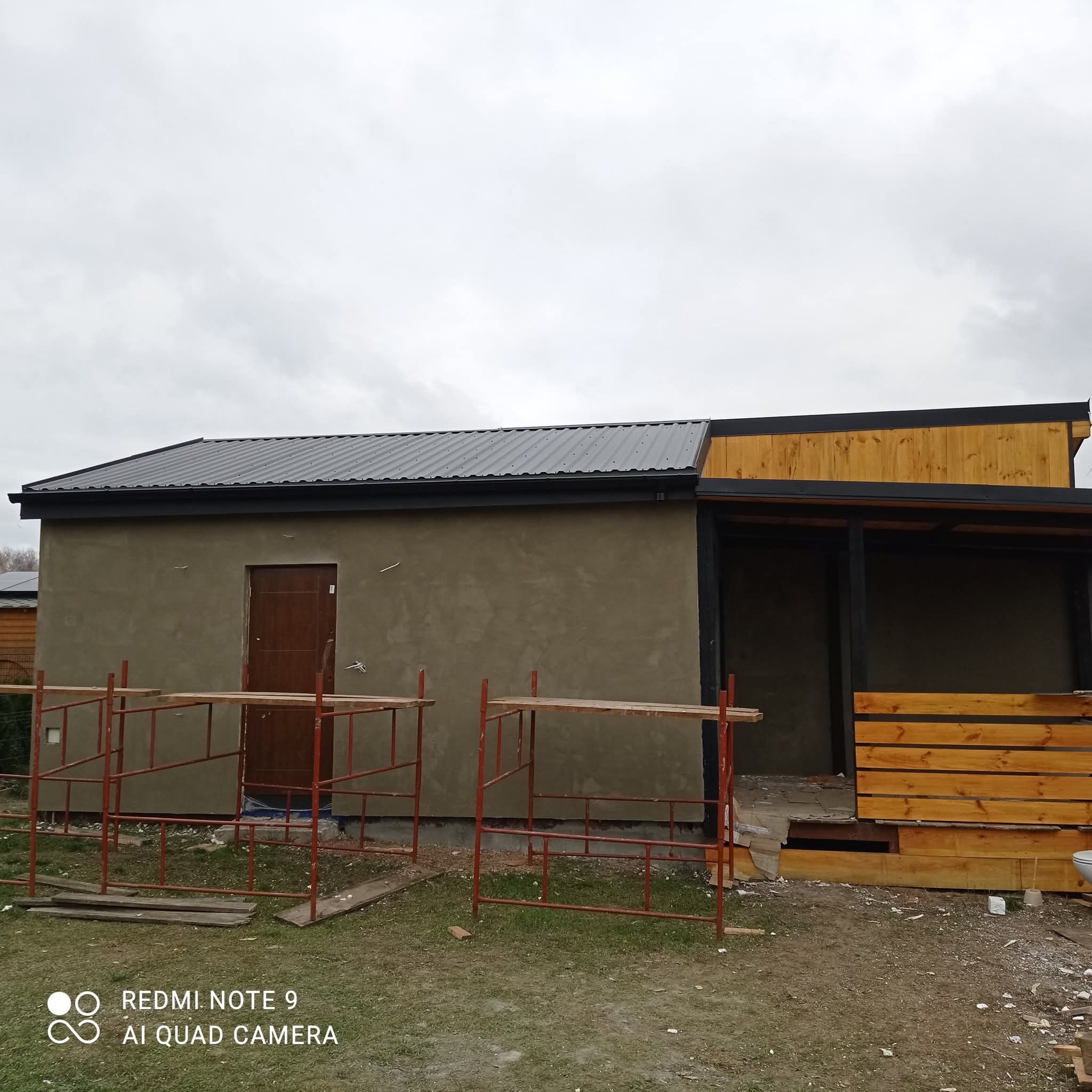 Budynek z szarą elewacją i ciemnym dachem z blachy falistej, w trakcie prac wykończeniowych. Widoczne rusztowania i drewniana konstrukcja werandy.