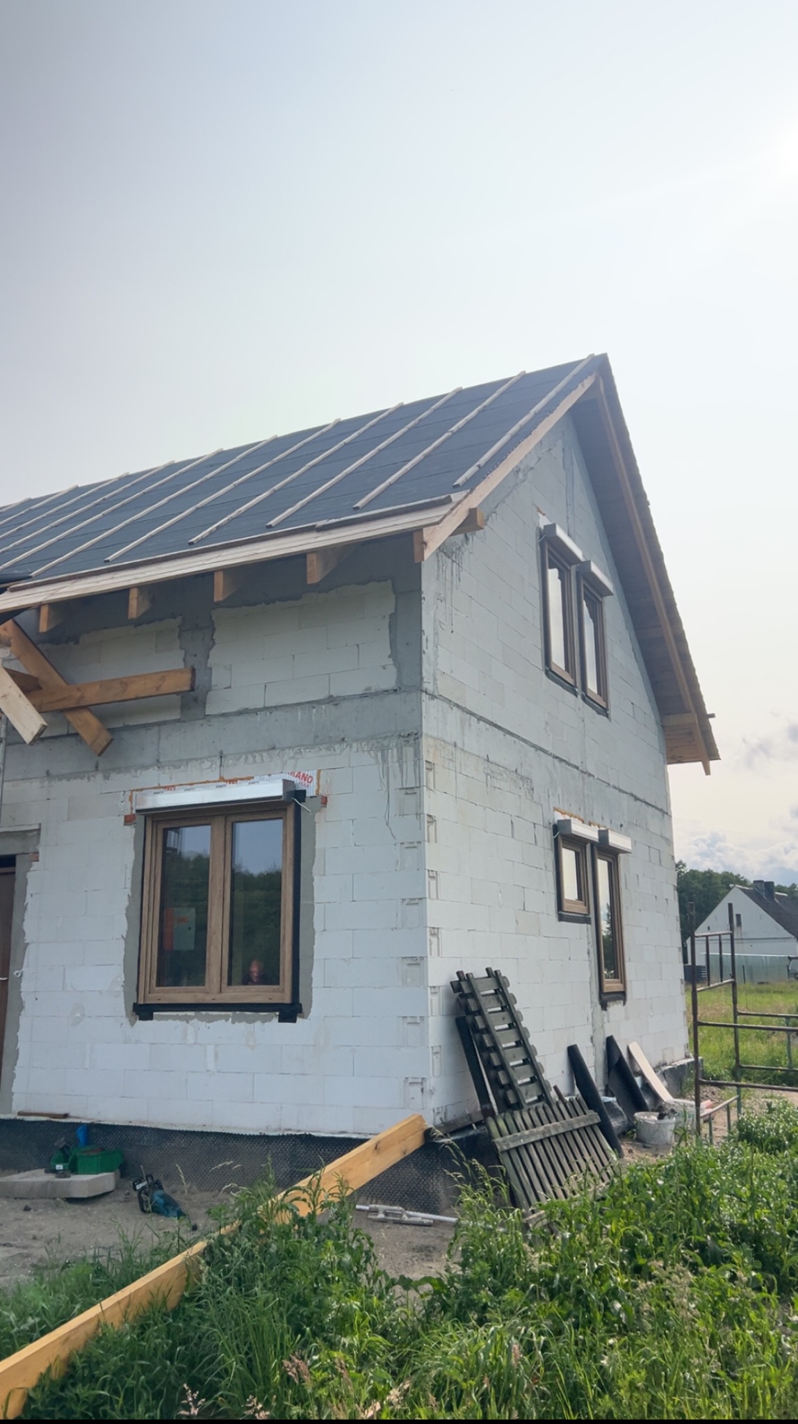 Budowa domu jednorodzinnego w trakcie krycia dachu, widoczne okna z roletami zewnętrznymi, ściany z białego bloczka. Częściowo położona dachówka.