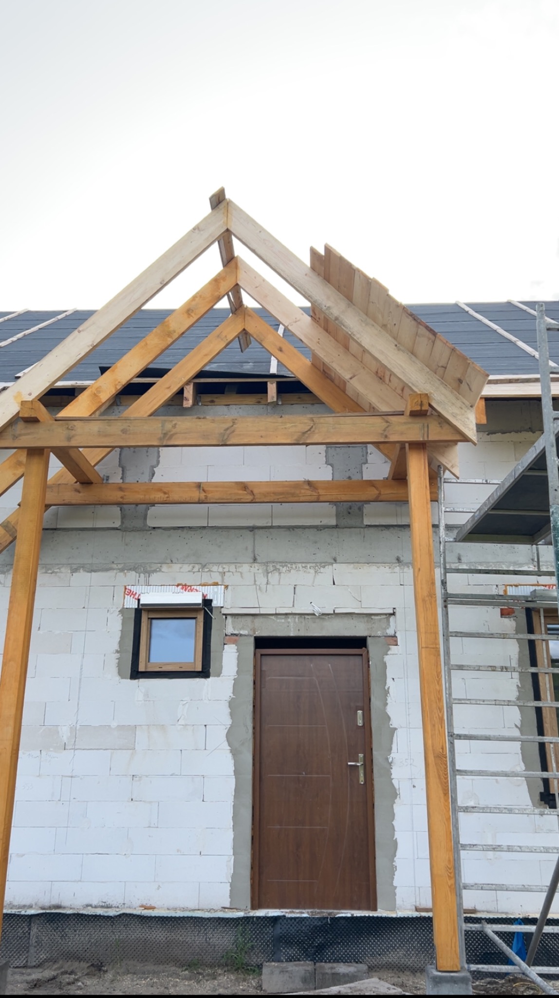 Budowa zadaszenia nad wejściem do domu z białego bloczka. Widoczna drewniana konstrukcja dachu i fragment pokrycia. Na ścianie okno i brązowe drzwi.