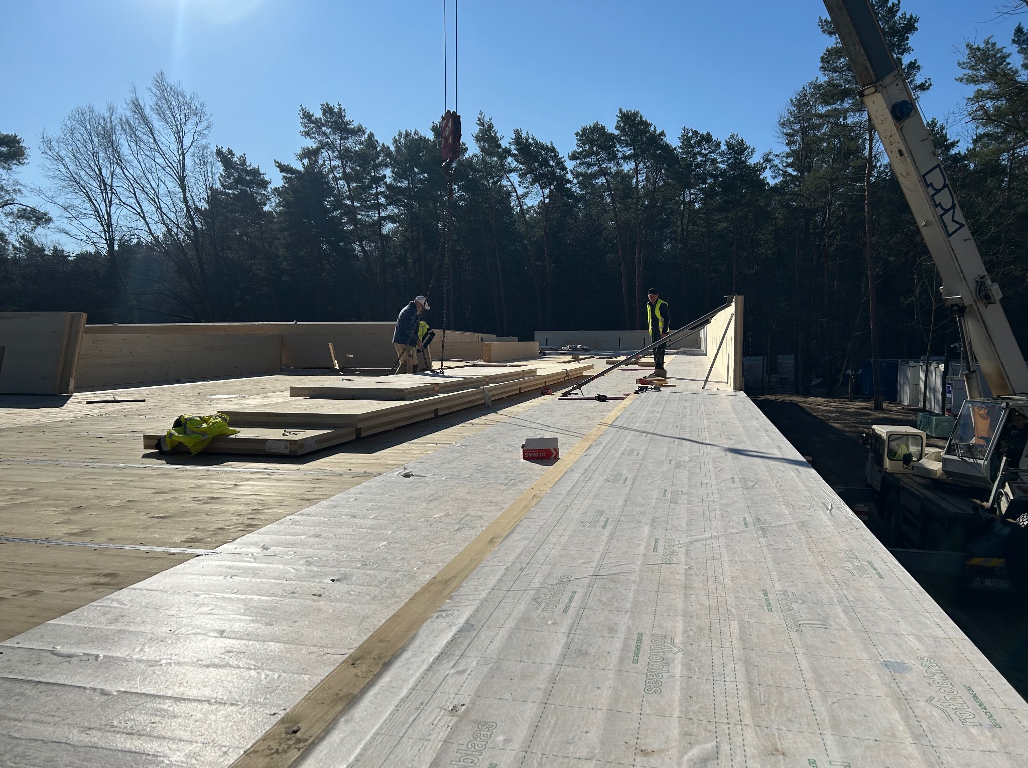 Montaż drewnianej konstrukcji dachu na budowie, widoczni pracownicy i dźwig. Elementy konstrukcyjne ułożone na przygotowanej powierzchni, las w tle, słoneczny dzień.