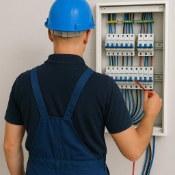 Adam Pałys - Elektryk w niebieskim kombinezonie sprawdza instalację elektryczną w rozdzielnicy, używając próbnika napięcia. Widok z tyłu, skupienie na tablicy z bezpiecznikami.