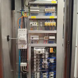 Otwarta szafa sterownicza z widocznymi elementami elektrycznymi: bezpieczniki, przekaźniki, okablowanie w kolorach. Widoczne oznaczenia napięć L1, L2, L3.