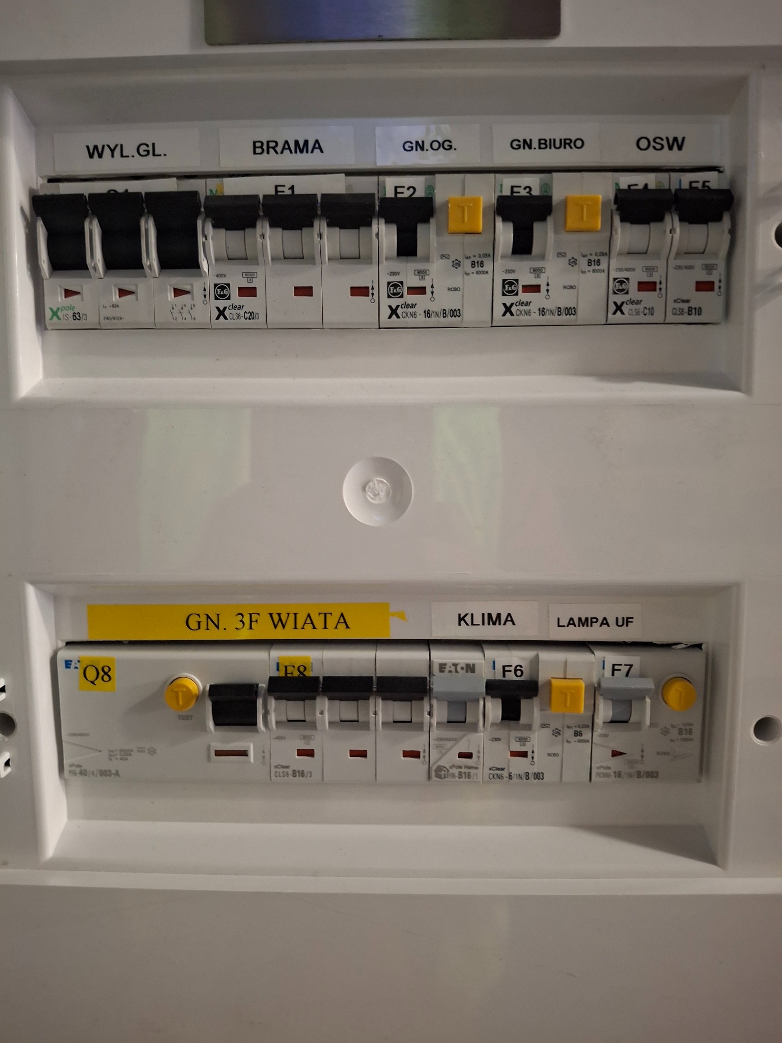 Rozdzielnia elektryczna z bezpiecznikami oznaczonymi etykietami: Wyl.Gl, Brama, Gn.Og, Gn.Biuro, OSW, Gn.3F Wiata, Klima, Lampa UF. Widoczne bezpieczniki i okablowanie.