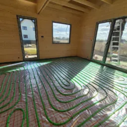 Jasne wnętrze domu w trakcie budowy z ułożonym ogrzewaniem podłogowym. Zielone rurki na folii, widok na okna i drewniane ściany.