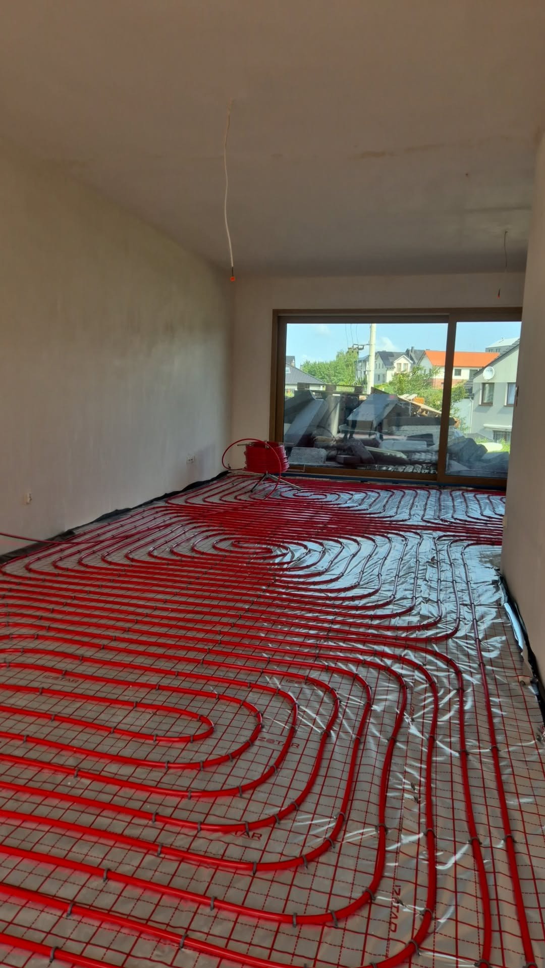 Czerwone rury ogrzewania podłogowego ułożone na folii w jasnym pokoju z dużym oknem. Widok na budynki za oknem. Etap instalacji przed wylewką.