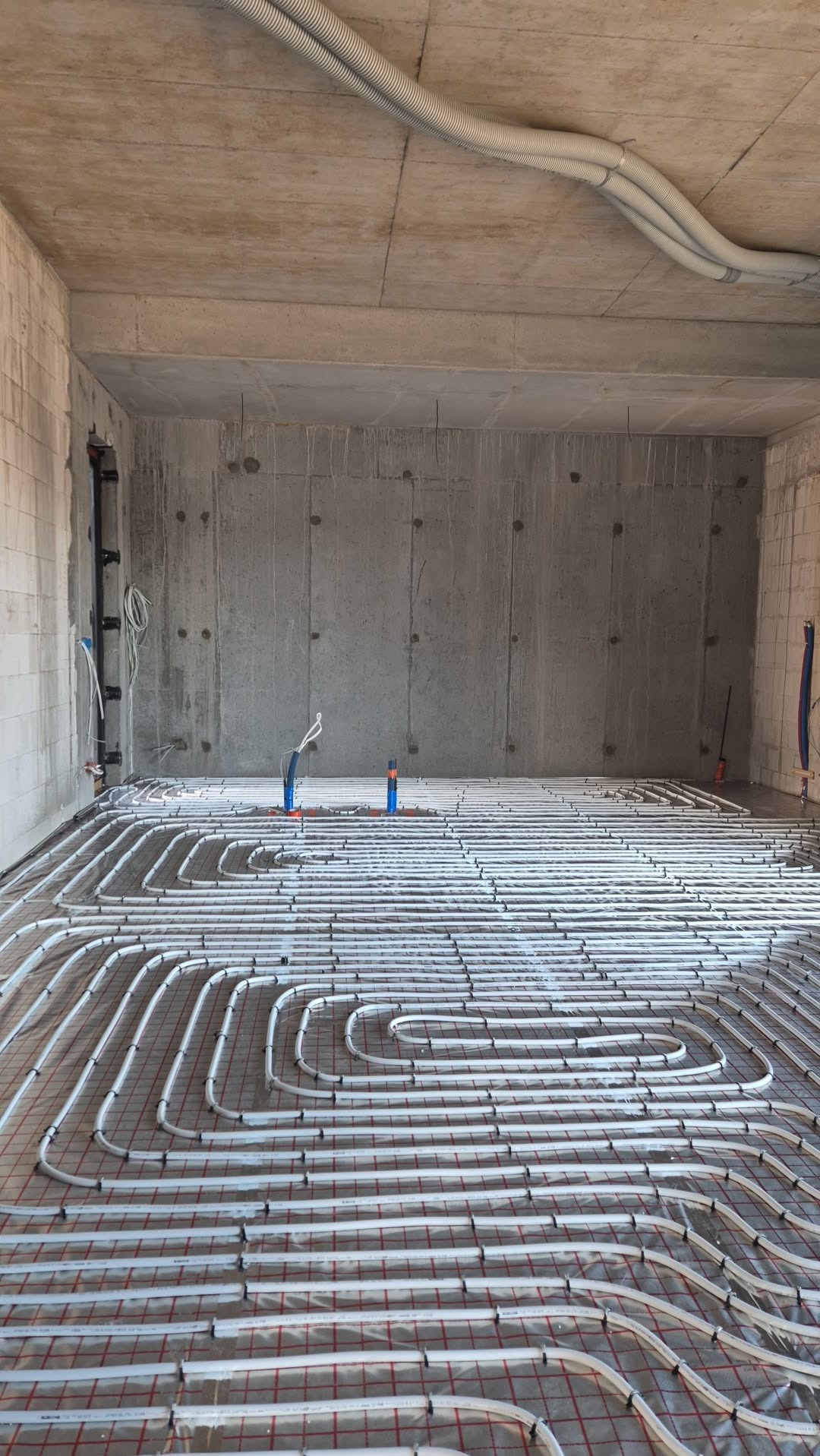 Białe rury ogrzewania podłogowego ułożone na folii z czerwoną siatką, w surowym wnętrzu z betonowymi ścianami i sufitem. Widoczne pionowe rury instalacyjne.