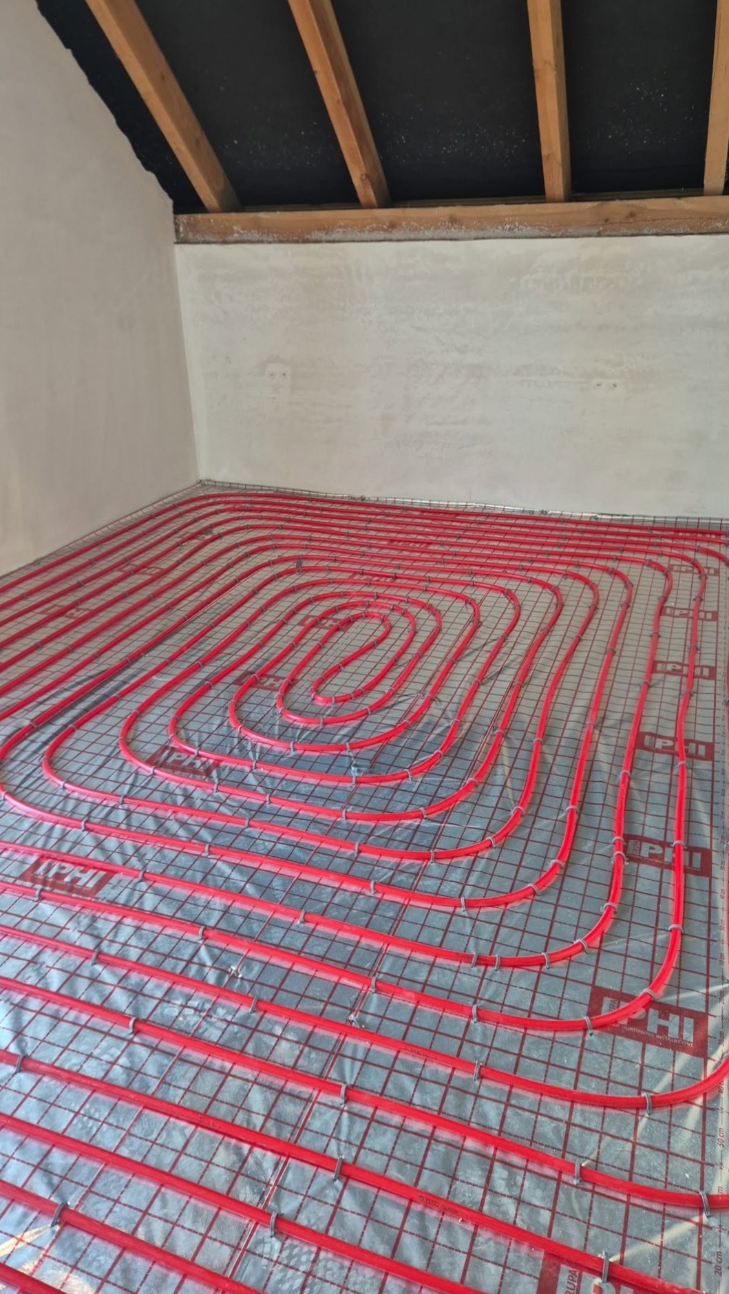 Czerwone rury ogrzewania podłogowego ułożone na srebrnej folii, widok z góry. Instalacja w trakcie realizacji w Kartuzach. Elementy mocujące widoczne.