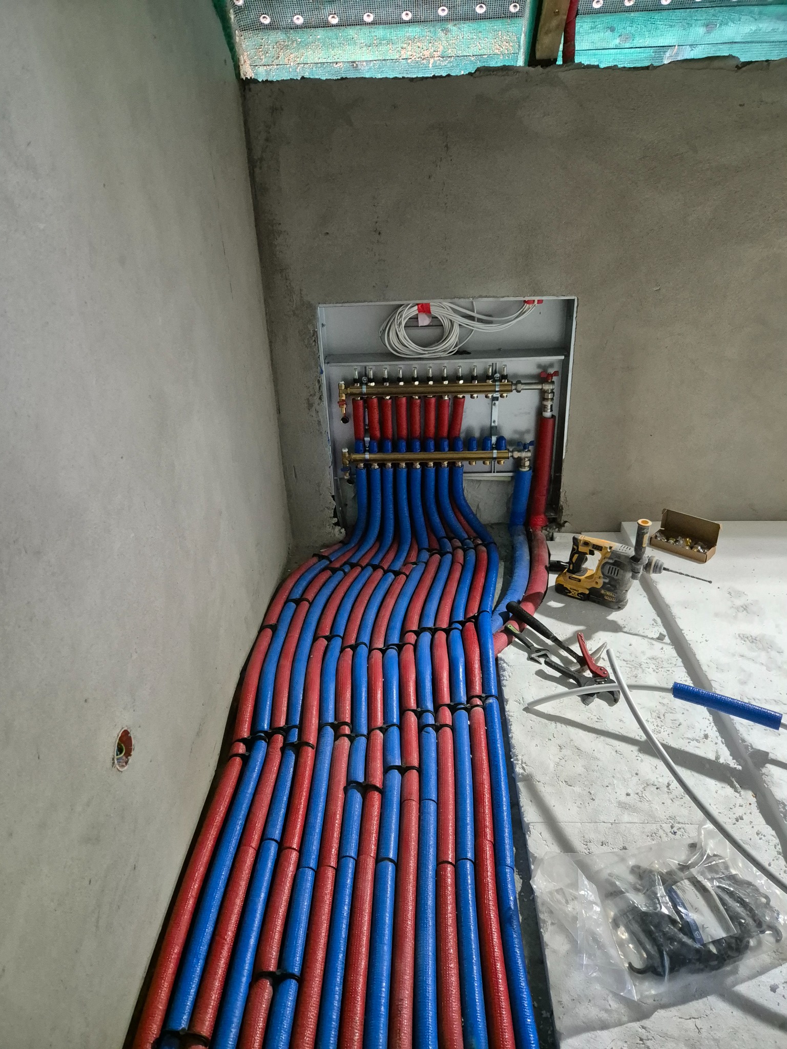 Rozłożone rury ogrzewania podłogowego (czerwone i niebieskie) prowadzące do rozdzielacza na tle surowych ścian. Widoczne narzędzia hydrauliczne i akcesoria montażowe.