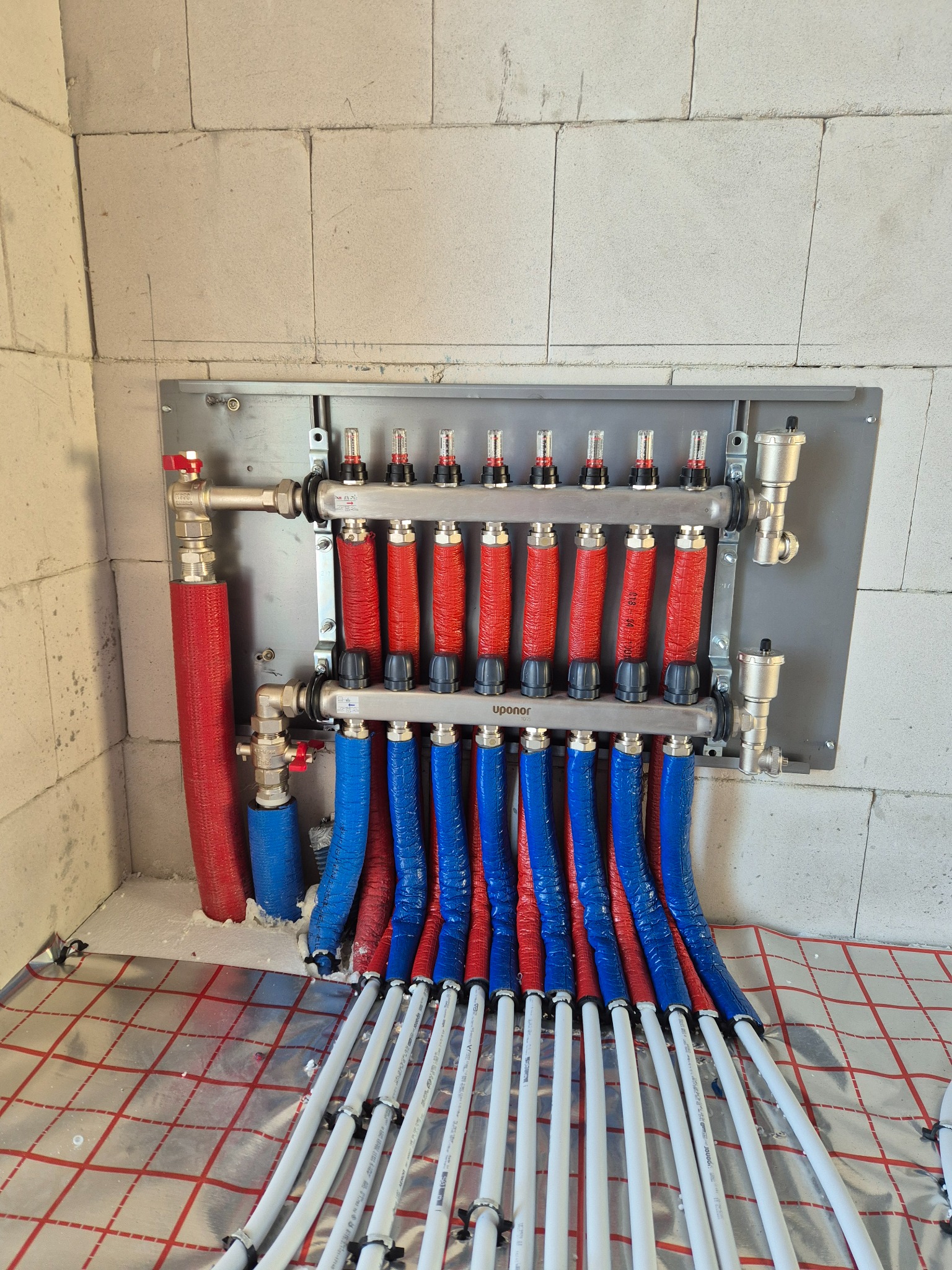 Rozdzielacz ogrzewania podłogowego Uponor z rurami w izolacji czerwonej i niebieskiej, zamontowany na ścianie z betonu komórkowego, widoczne białe rury na folii grzewczej.