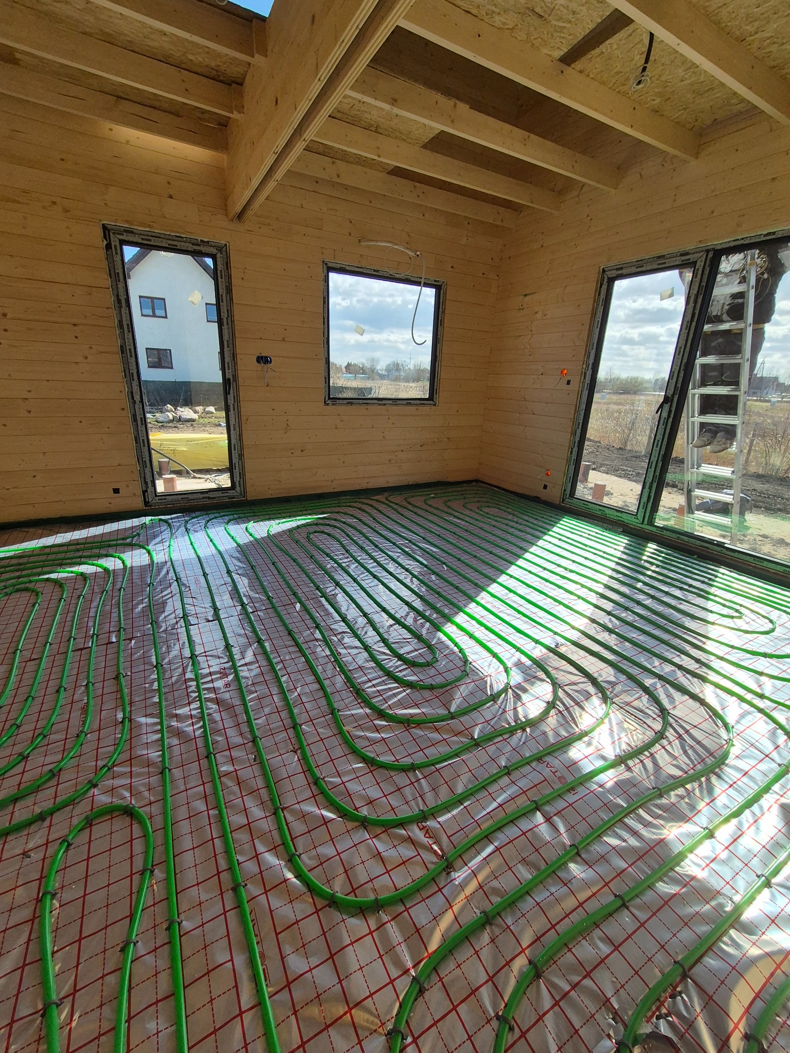 Jasne wnętrze domu w trakcie budowy z ułożonym ogrzewaniem podłogowym. Zielone rurki na folii, widok na okna i drewniane ściany.