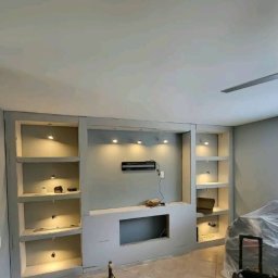 Kompleksowe Remonty - Ściana działowa z regipsu z wnękami na półki i TV w szarym odcieniu, oświetlenie punktowe, w trakcie prac wykończeniowych. Widoczne narzędzia pracy.