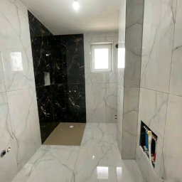 Kompleksowe Remonty - Wykończona łazienka z czarno-białymi płytkami imitującymi marmur, widoczny brodzik i okno. W ścianie otwór na instalacje. Całość w jasnej tonacji.