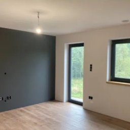 Kompleksowe Remonty - Wnętrze pokoju po malowaniu: jedna ściana grafitowa, reszta biała. Widoczne okno z czarną ramą, drewniana podłoga i przygotowane punkty elektryczne.