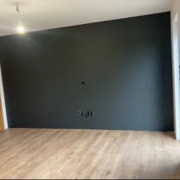 Kompleksowe Remonty - Pomalowane na ciemny kolor ściany w pokoju z drewnianą podłogą i drzwiami. Widoczne gniazdka elektryczne na ścianie i wiszący kabel od lampy na suficie.