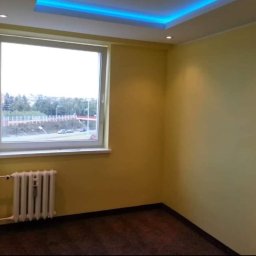 Kompleksowe Remonty - Pomalowany pokój z oknem i grzejnikiem, ściany w odcieniach żółci, brązowa podłoga, sufit z podświetleniem LED. Widok z okna na zieleń i most.